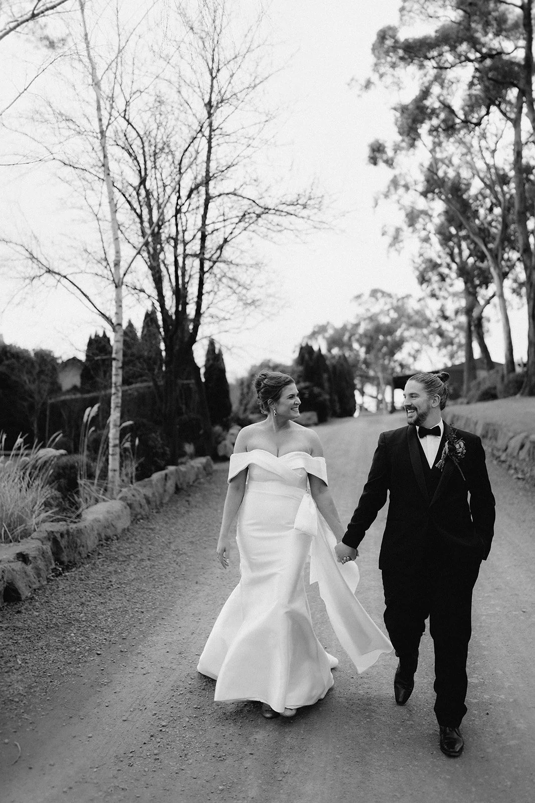 A black-and-white photo of a bride and groom walking hand in hand outdoors on a dirt path, surrounded by trees and bushes. The bride is in a strapless wedding gown, and the groom is in a black tuxedo with a bow tie.