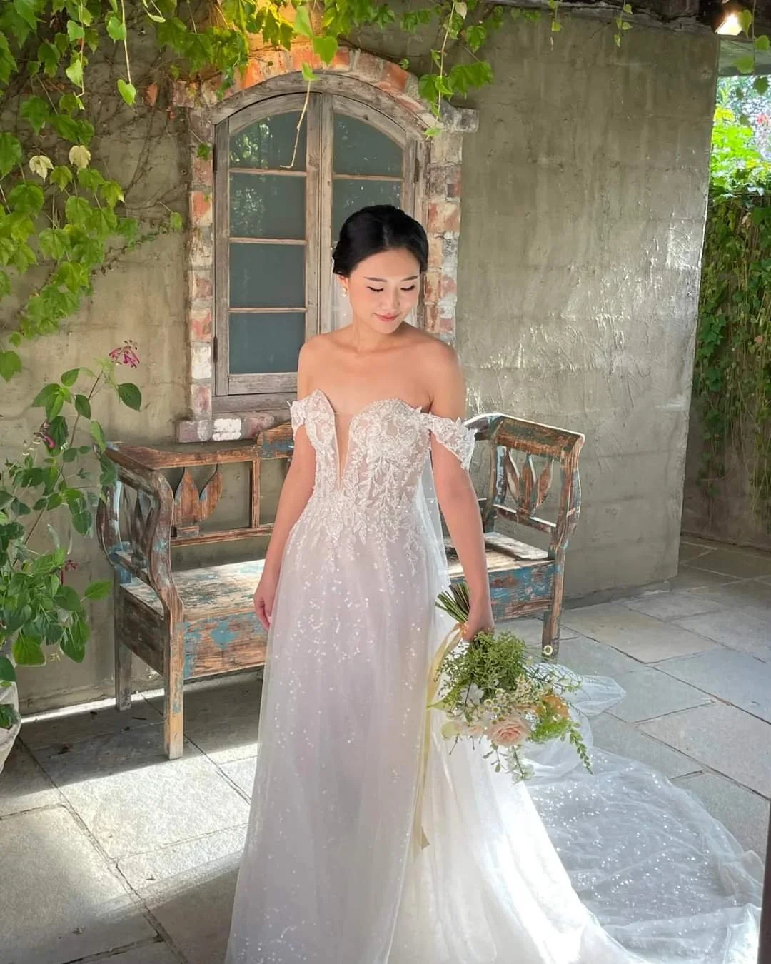 A bride in a white wedding dress holding a bouquet of flowers, standing outdoors near a rustic stone and brick wall with an old window and green vines.