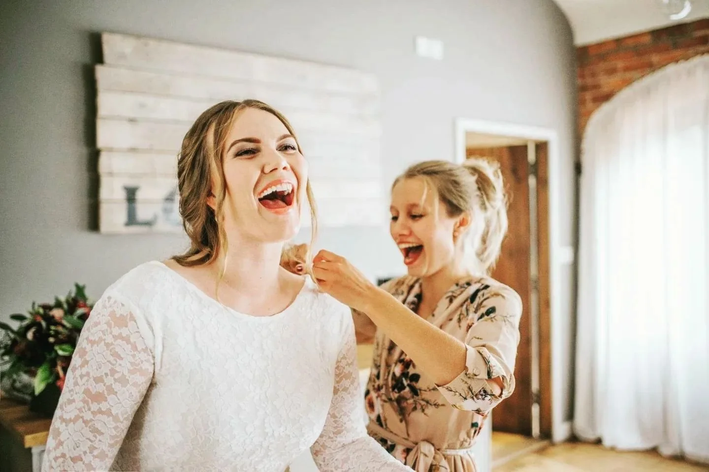 Two women smiling and laughing, one helping the other put on a lace wedding dress in a bright room with white curtains and wooden accents.