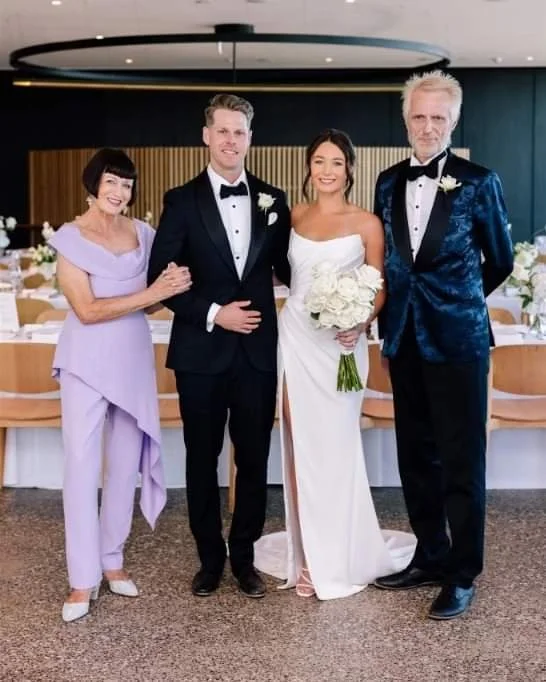 A group of four people, including a bride in a white wedding dress holding a bouquet, standing with a groom in a black tuxedo, and two older adults, all dressed formally, inside a decorated event space with round tables and floral arrangements.