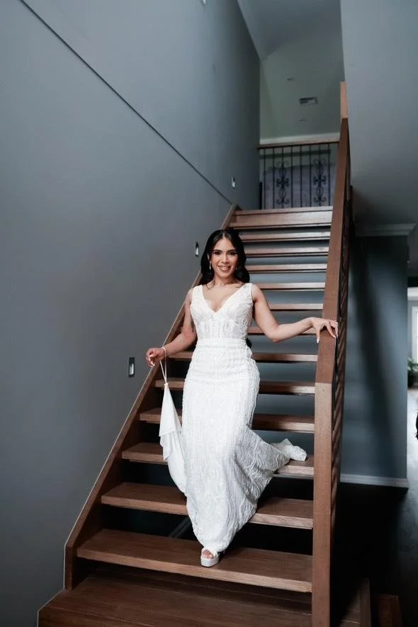 Bride in a fitted beaded white wedding gown standing on a wooden staircase inside a house.