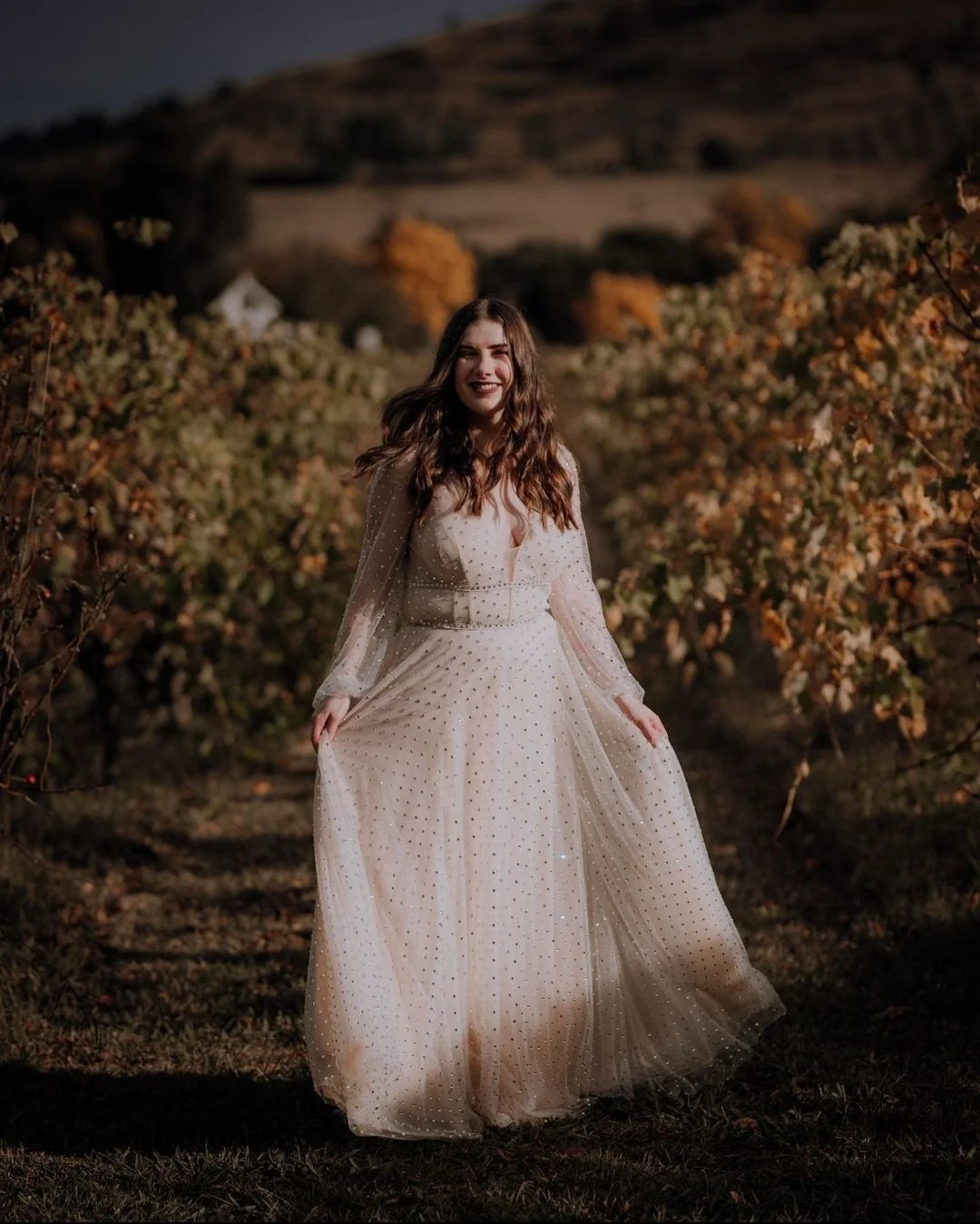 A young woman in an elegant, long white dress with sparkling embellishments stands in a vineyard during dusk, smiling happily.