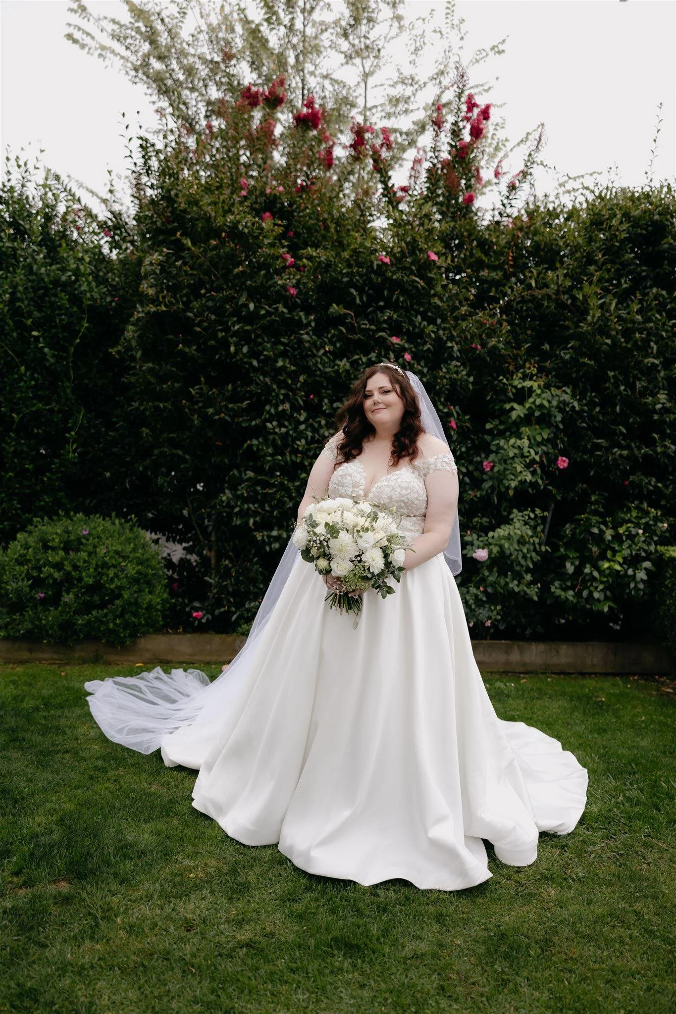 Bride in a wedding gown holding a bouquet of white flowers standing on a green lawn with bushes and flowering plants in the background.