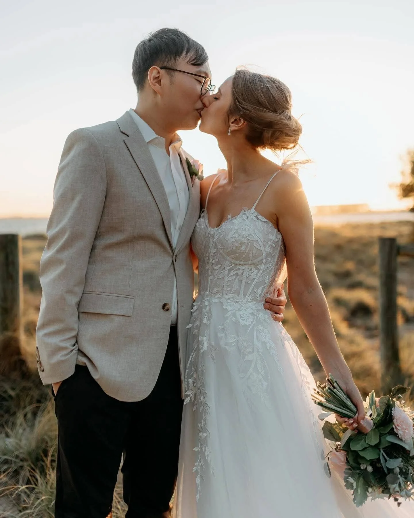 A newlywed couple sharing a kiss outdoors at sunset, the bride in a lace wedding dress holding a bouquet, and the groom in a light gray suit with glasses.