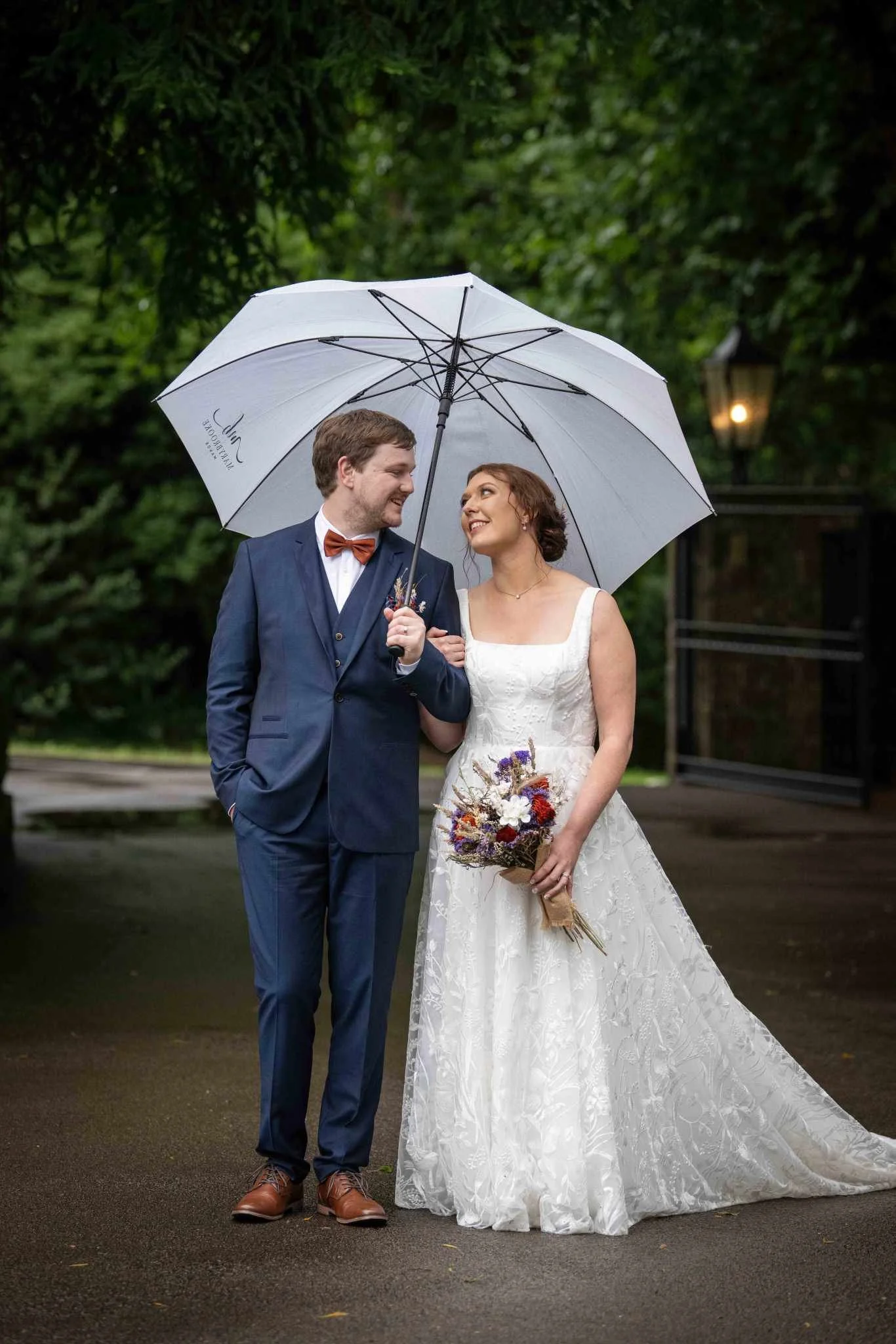 A bride and groom standing together under an umbrella outdoors on a rainy day, with trees and a lamppost in the background. The groom is wearing a dark suit with a bow tie, and the bride is wearing a white lace wedding dress, holding a bouquet of flo