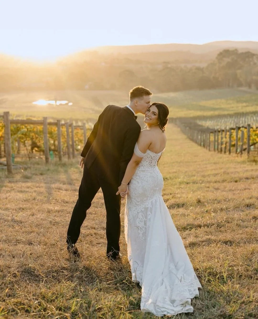 A newlywed couple in wedding attire holding hands and sharing a kiss in a vineyard at sunset.