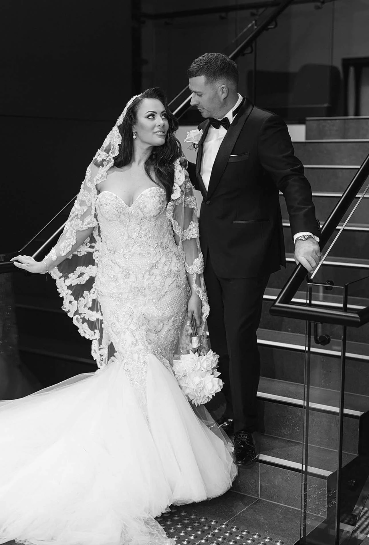 Black and white photo of a bride and groom on stairs, looking at each other, with the bride holding a bouquet of flowers.