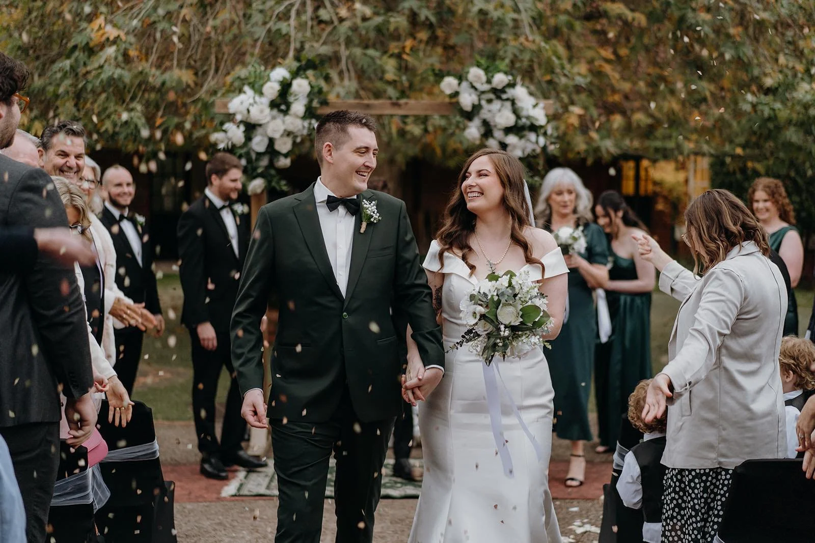 A newly married couple holding hands smiling at each other during their outdoor wedding ceremony. The groom is in a black tuxedo and the bride in a white wedding dress holding a bouquet. Guests and wedding party are celebrating around them.