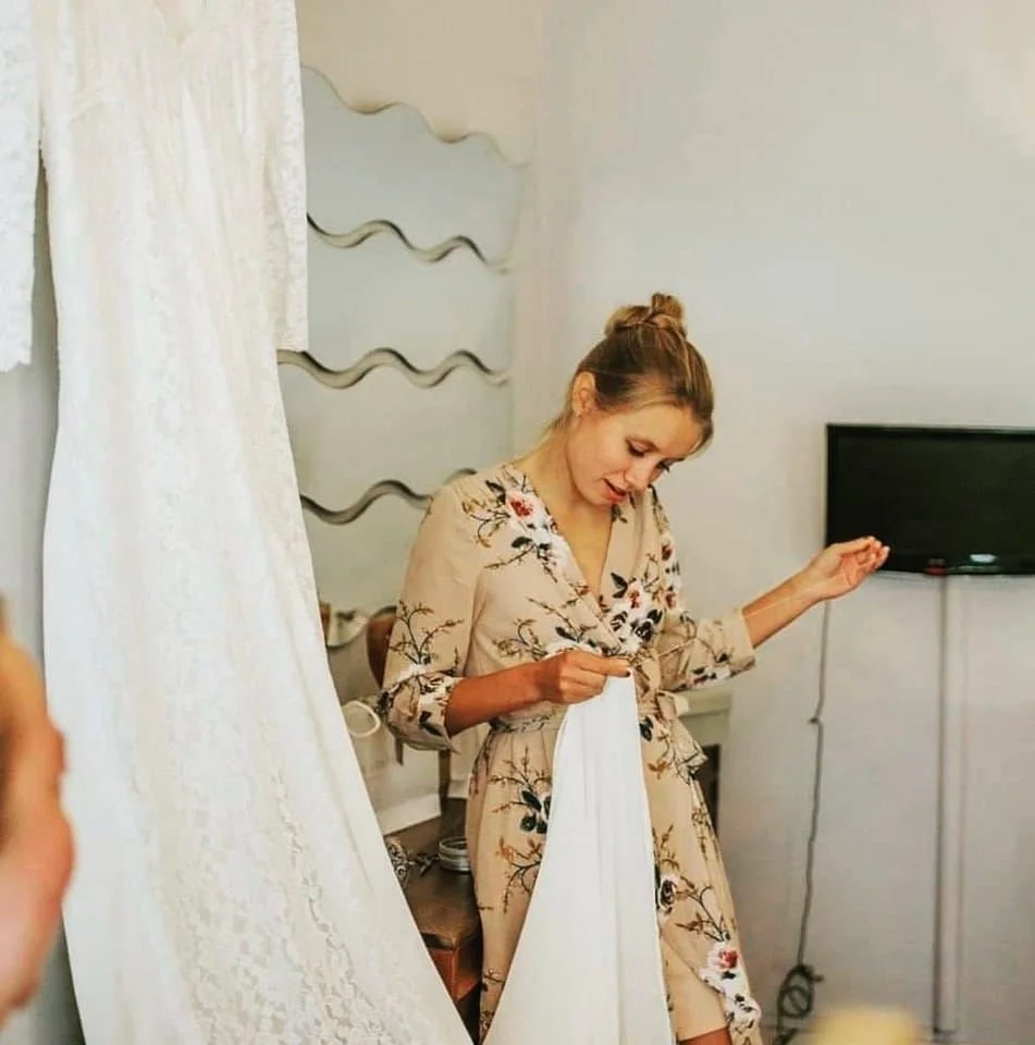 A woman with her hair in a bun, wearing a beige floral dress, standing in a room with a white curtain, a TV on the wall, and a decorative metal wall art behind her, hand sewn a wedding dress.