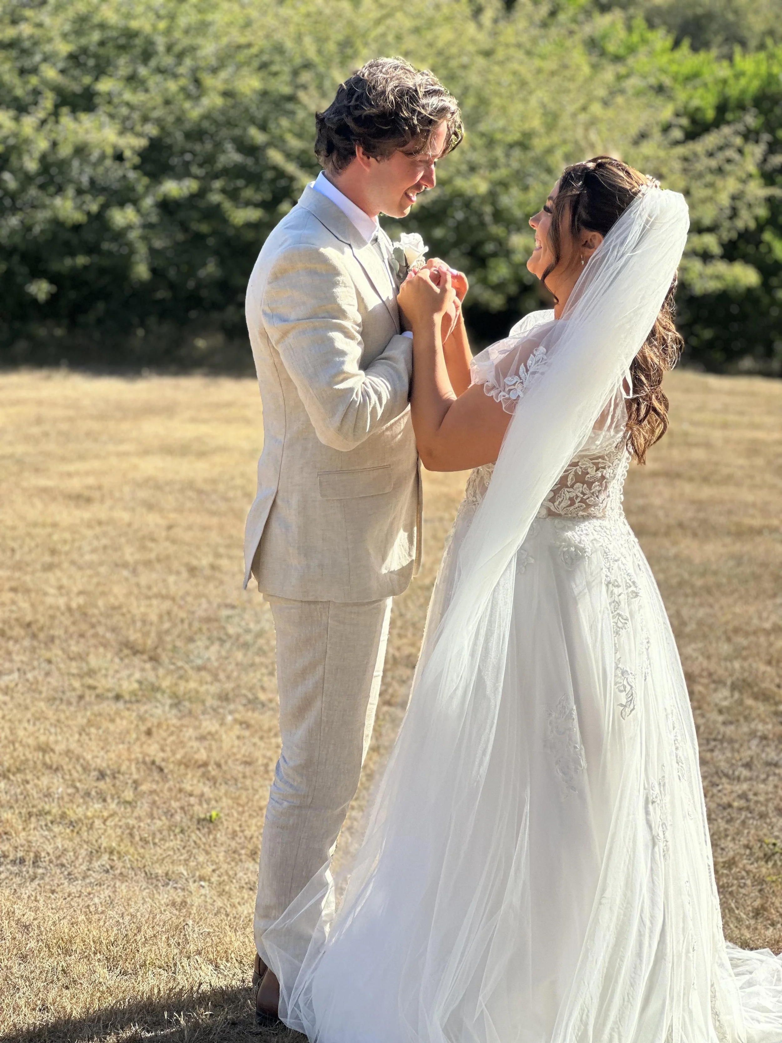 A bride and groom holding hands and smiling at each other outdoors on a sunny day, with green trees in the background. The bride is wearing a white lace wedding gown and veil, and the groom is in a light-colored suit.