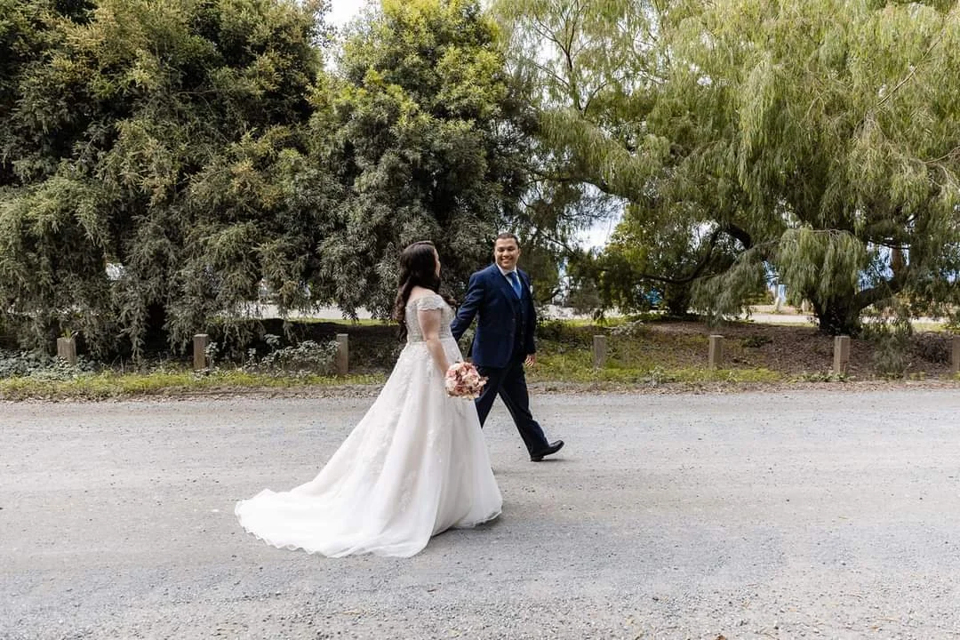 A bride and groom walking outdoors on a gravel path, holding hands, with trees and greenery in the background. The bride is wearing a white wedding dress and holding a bouquet of flowers, while the groom is dressed in a dark suit.
