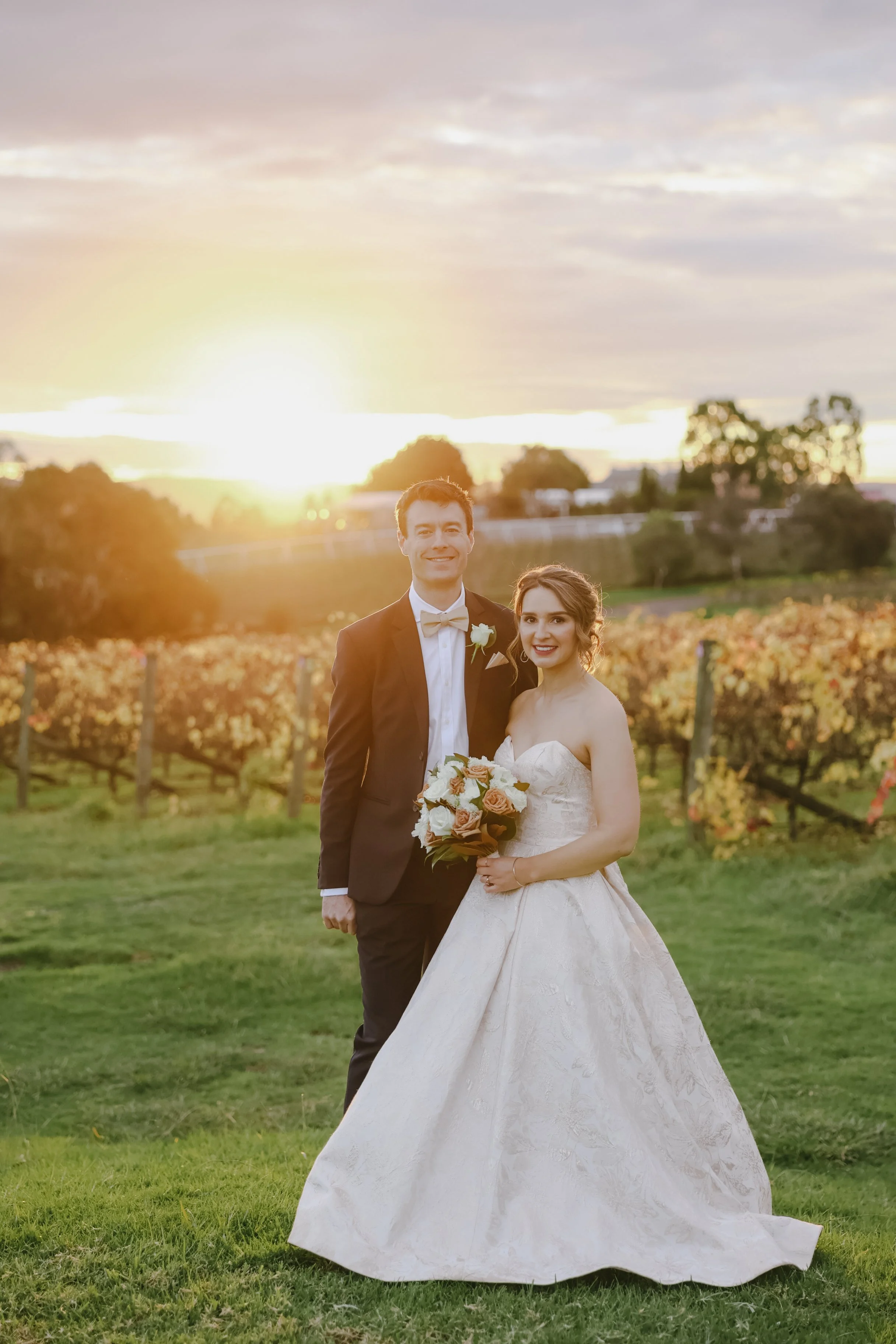 A newlywed couple standing in a vineyard during sunset. The groom in a black tuxedo and bowtie, and the bride in a white wedding gown holding a bouquet of flowers, smiling at the camera.