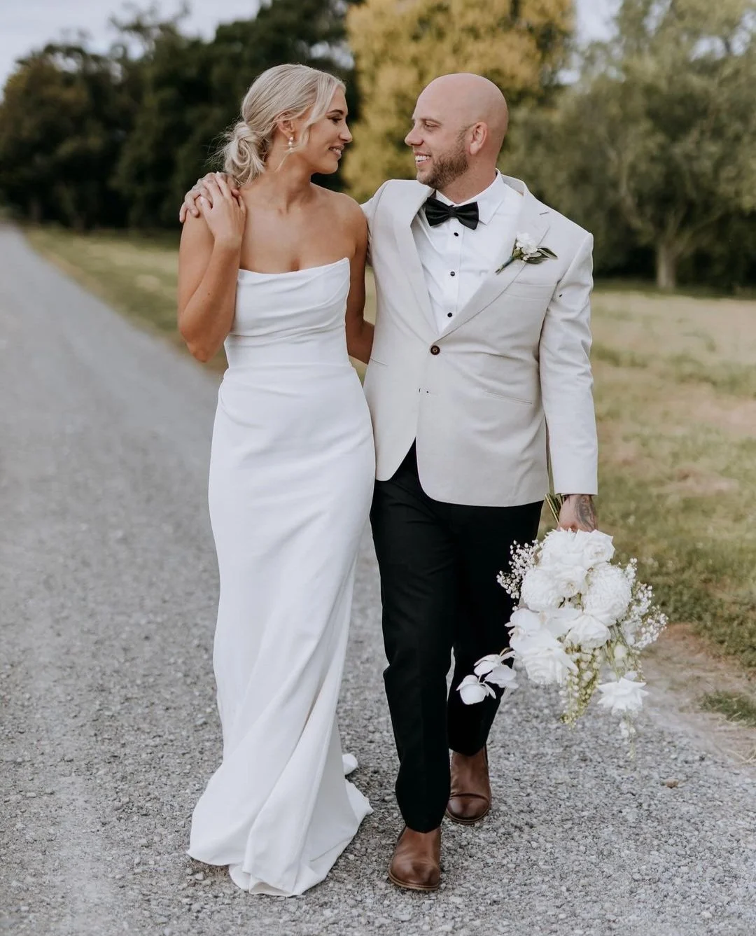 A bride and groom walking outdoors, smiling at each other. The bride wears a 2nd hand strapless ivory fitted wedding gown from KYHA, and the groom is in a light suit with a black bow tie, holding a bouquet of white flowers.