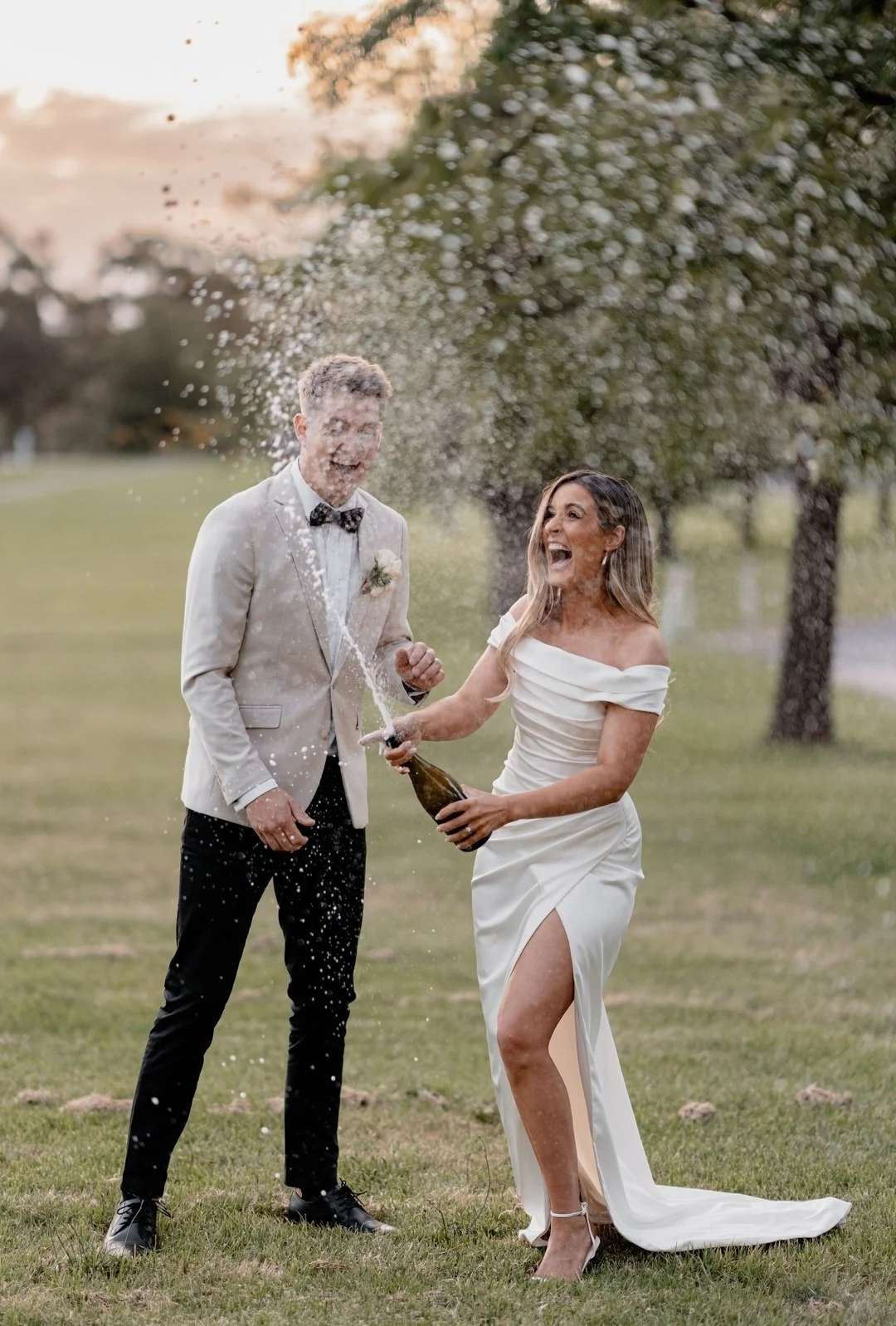 A newlywed couple in wedding attire celebrating outdoors by opening a champagne bottle, with champagne spraying out as they laugh and smile.