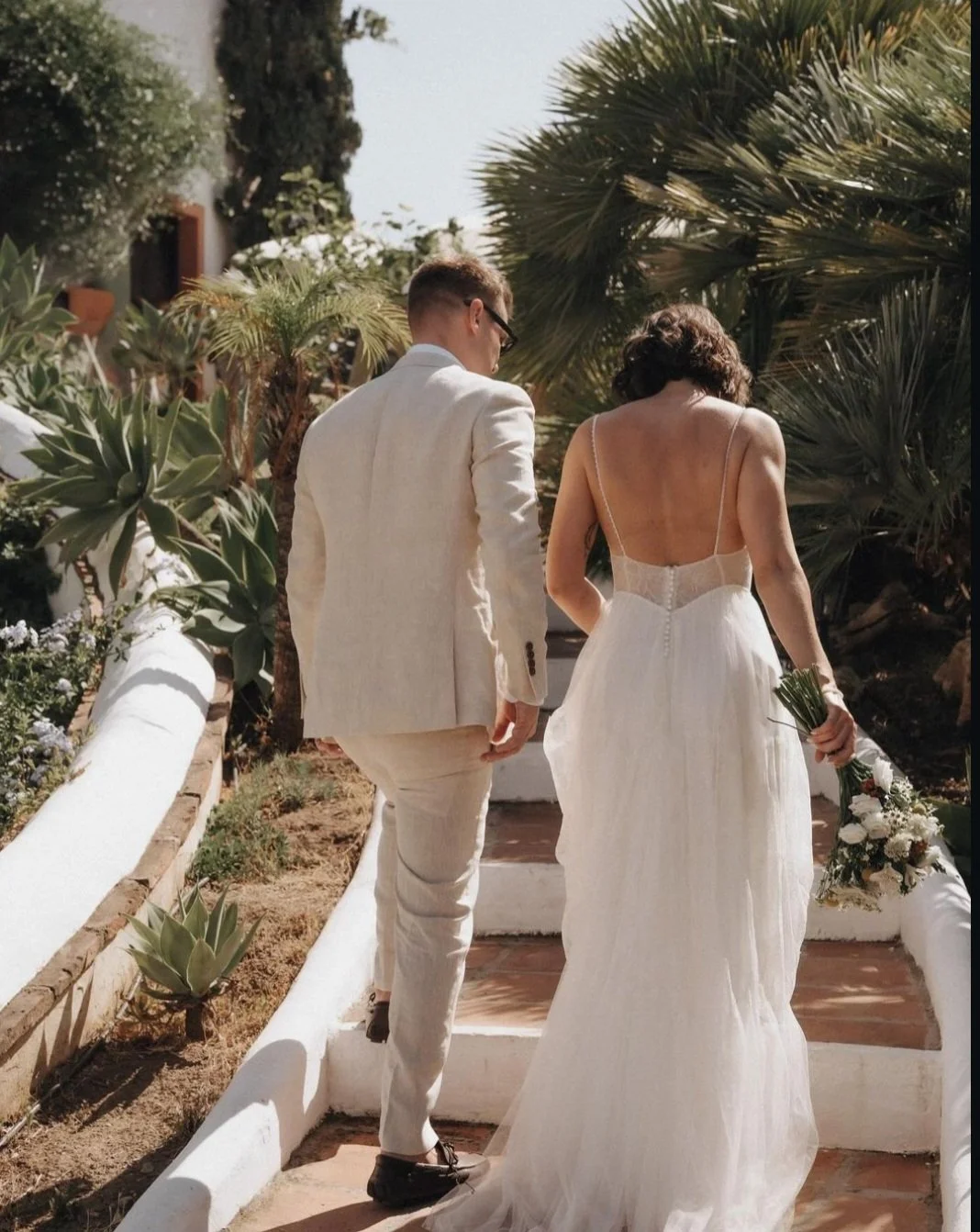 Bride in a tailored wedding gown and groom walking up garden steps with lush greenery, bride holding a bouquet after expert bridal alterations in Melbourne.