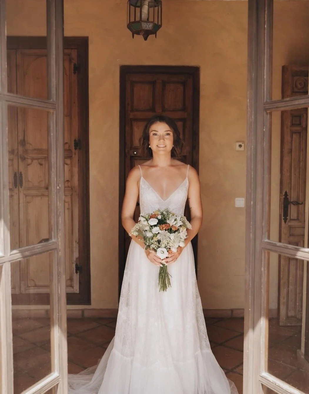 Bride in a resized and restyled white wedding gown holding a bouquet of flowers, standing in a rustic room with wooden doors, after expert bridal alterations in Melbourne