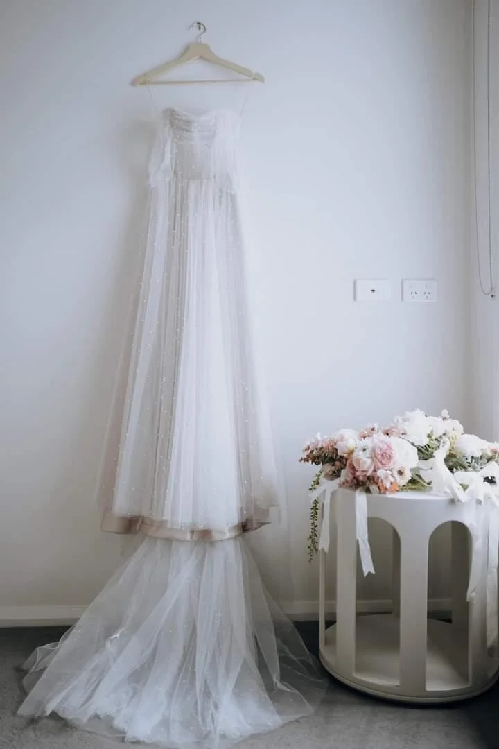 A white wedding dress hanging on a hanger against a plain wall, with a bouquet of pink and white flowers on a small white round table nearby.