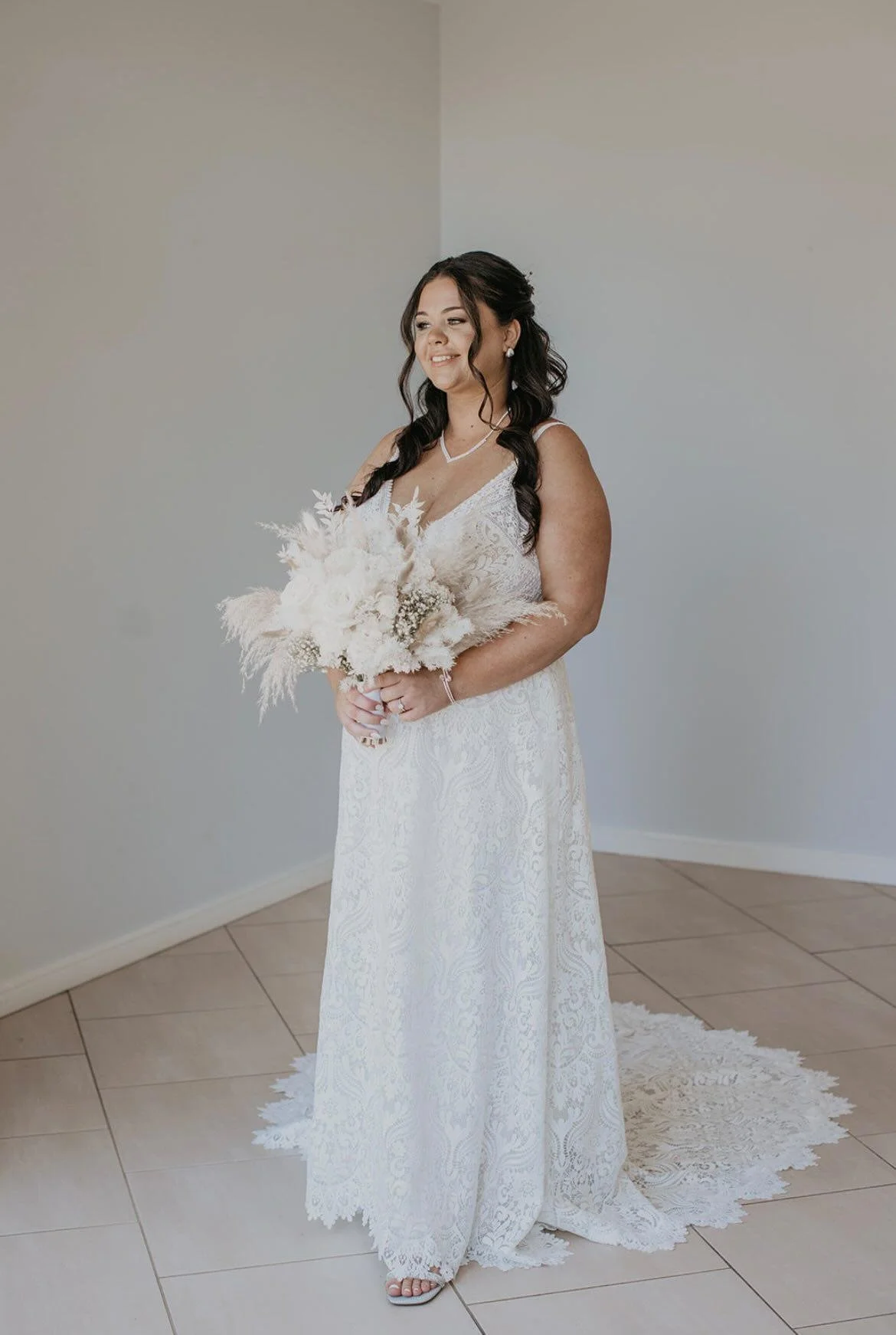 A woman in a white lace wedding dress holding a bouquet of dried flowers, standing on a tiled floor with a white lace rug behind her, in a minimal room with light-colored walls.