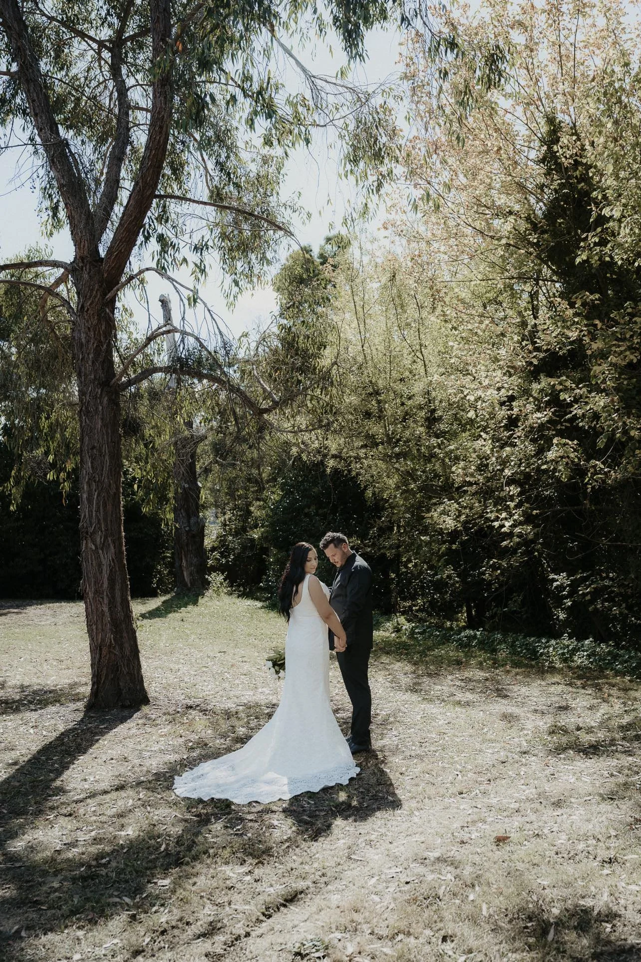 A bride and groom stand close together holding hands in a wooded outdoor area on their wedding day, surrounded by trees and greenery.