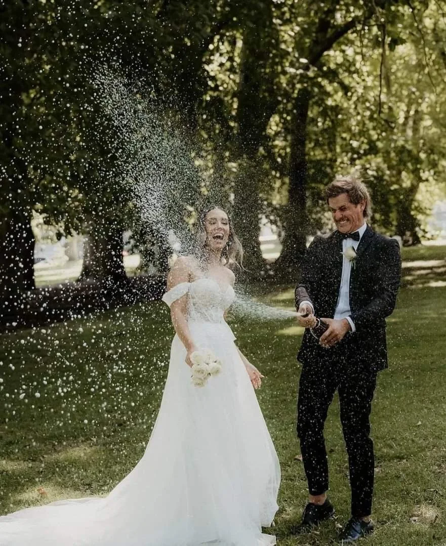 A newlywed couple celebrating outdoors with champagne and a water spray, with trees and sunlight in the background.
