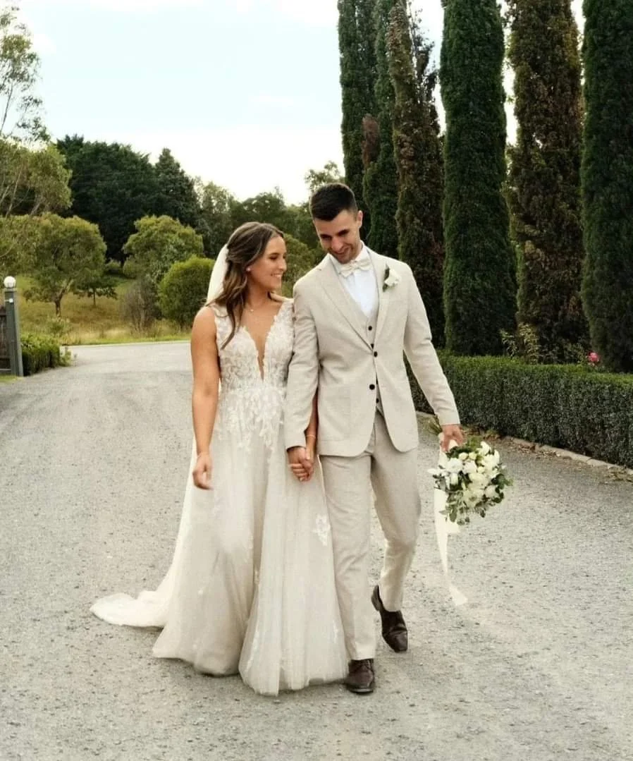 A bride and groom walking hand-in-hand outdoors on their wedding day, smiling and looking at each other, with lush greenery and tall trees in the background.