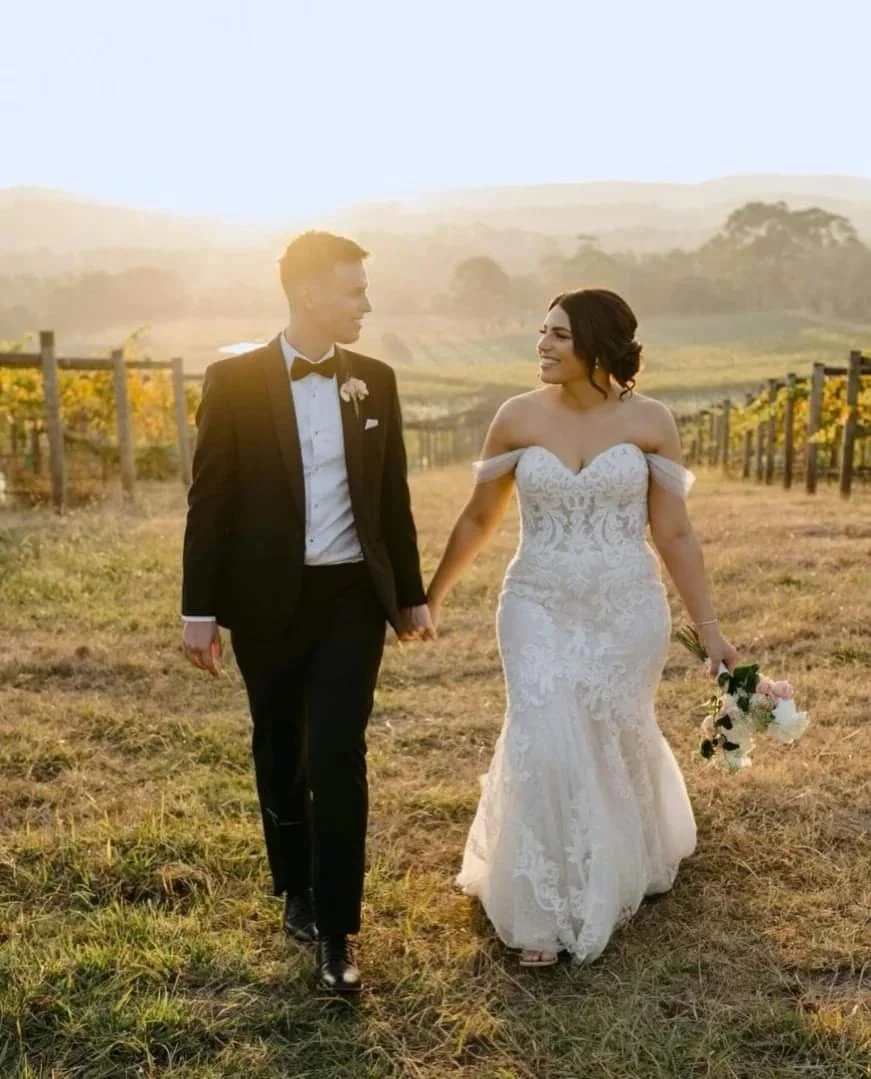 A bride and groom walk hand in hand through a vineyard at sunset, with the bride holding a bouquet and both dressed in wedding attire.