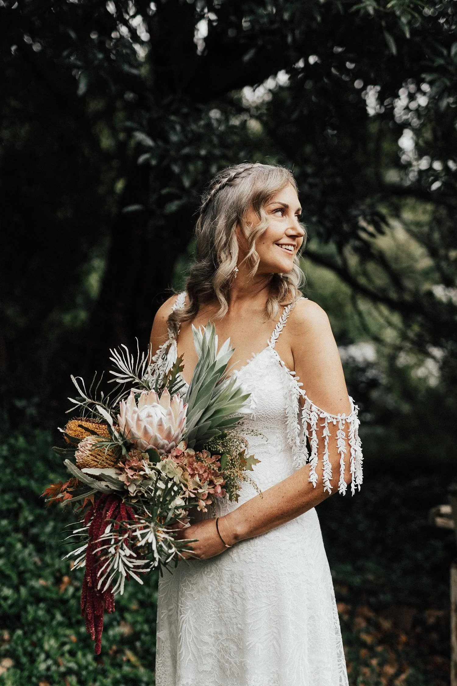 A bride holding a bouquet of tropical flowers and greenery, standing outdoors in a forested area, smiling and looking to her left.