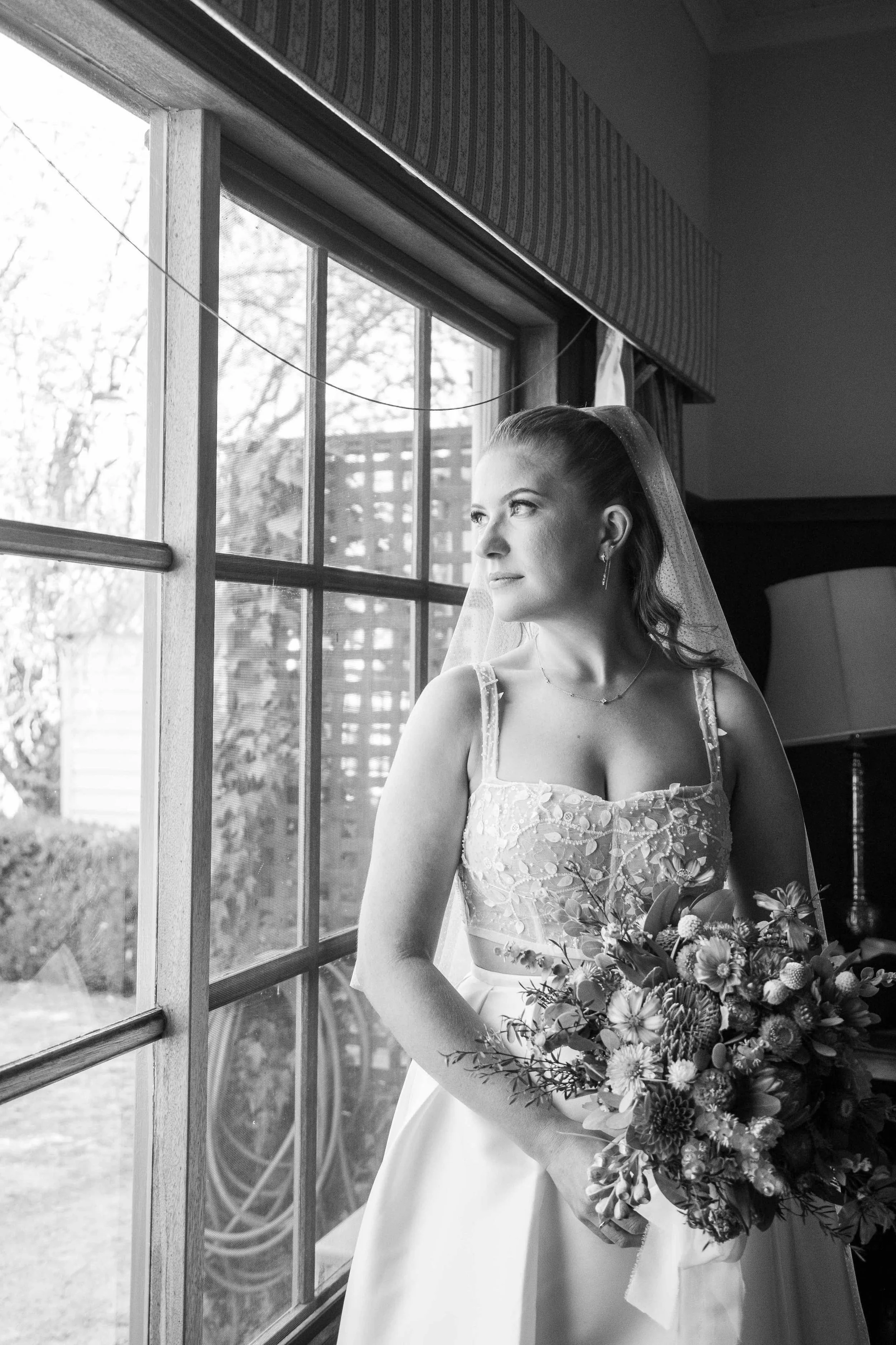 Black and white photo of a bride in a custom beaded corset standing by a window, holding a bouquet of flowers, looking out thoughtfully.