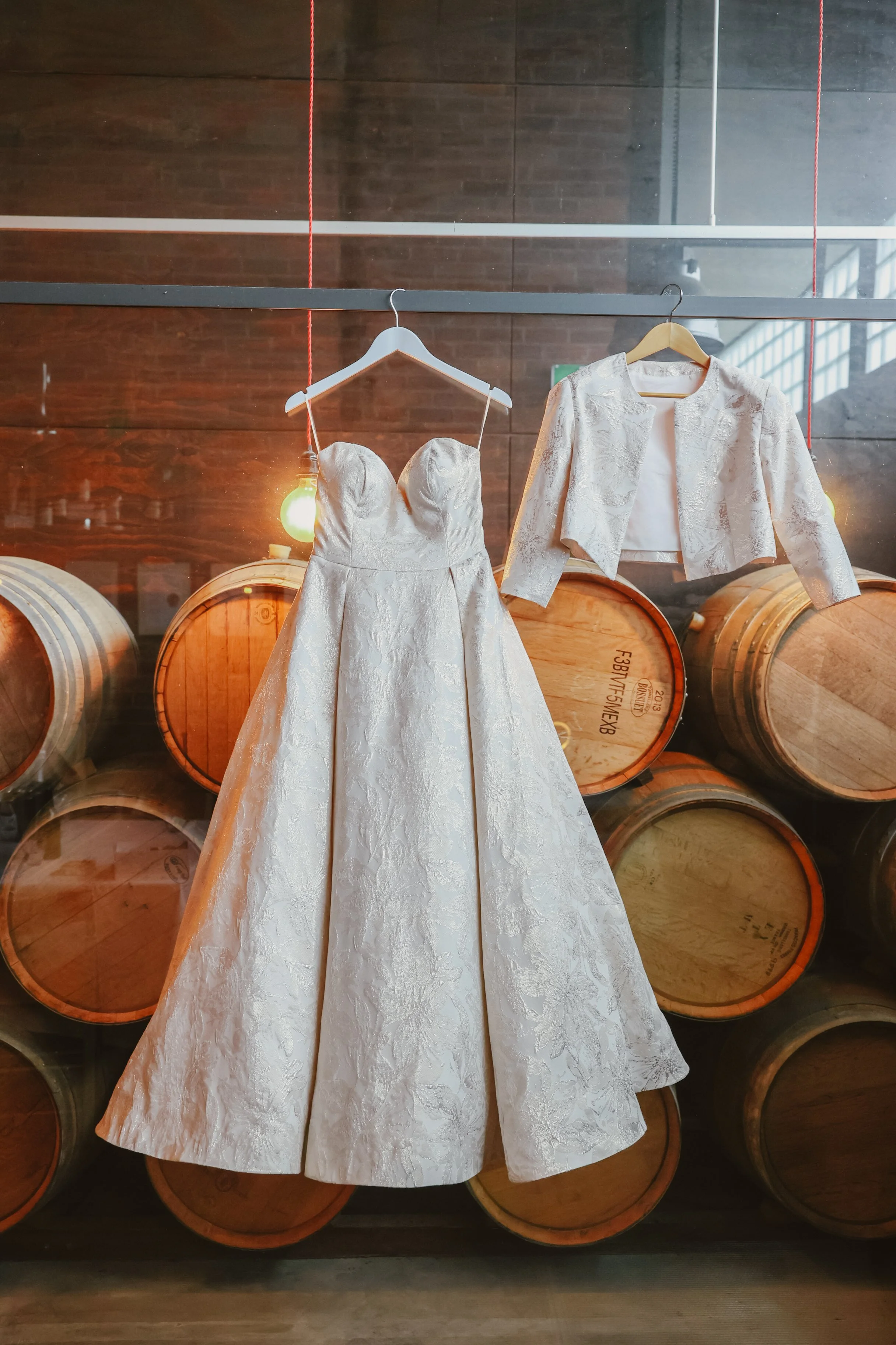 A white wedding dress and a matching jacket hanging on a rack, displayed in front of wooden wine barrels.