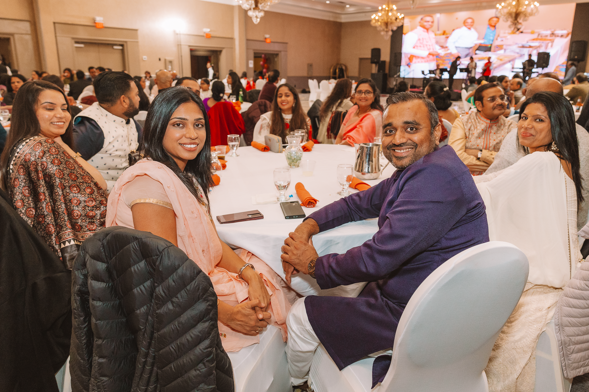 A diverse group of people sitting around a banquet table at an event, with some smiling at the camera. The background shows a stage with a large screen and musical instruments, indicating a performance or presentation is taking place. The attendees are dressed in semi-formal attire.