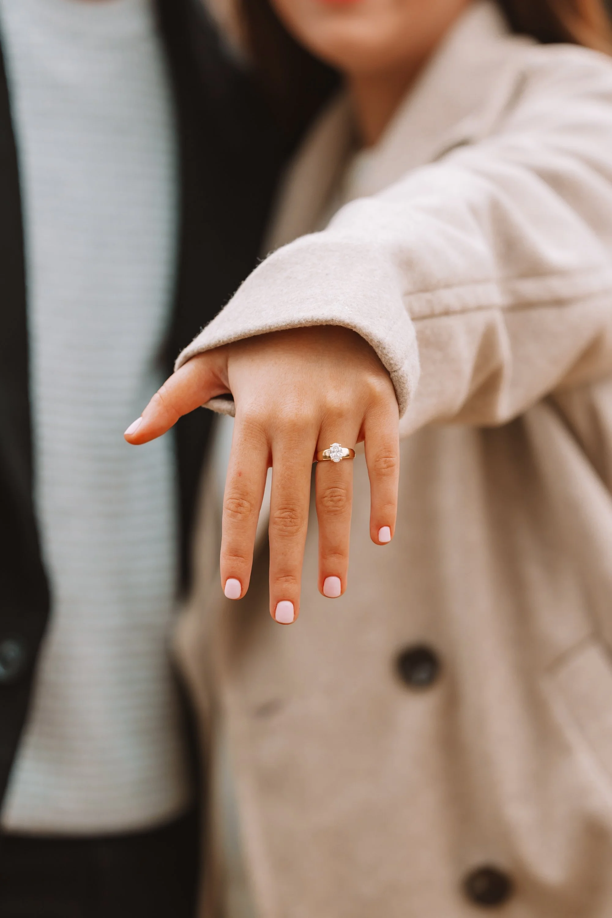 A person showing their left hand with a diamond engagement ring on the ring finger, wearing a beige coat with a rolled-up sleeve.