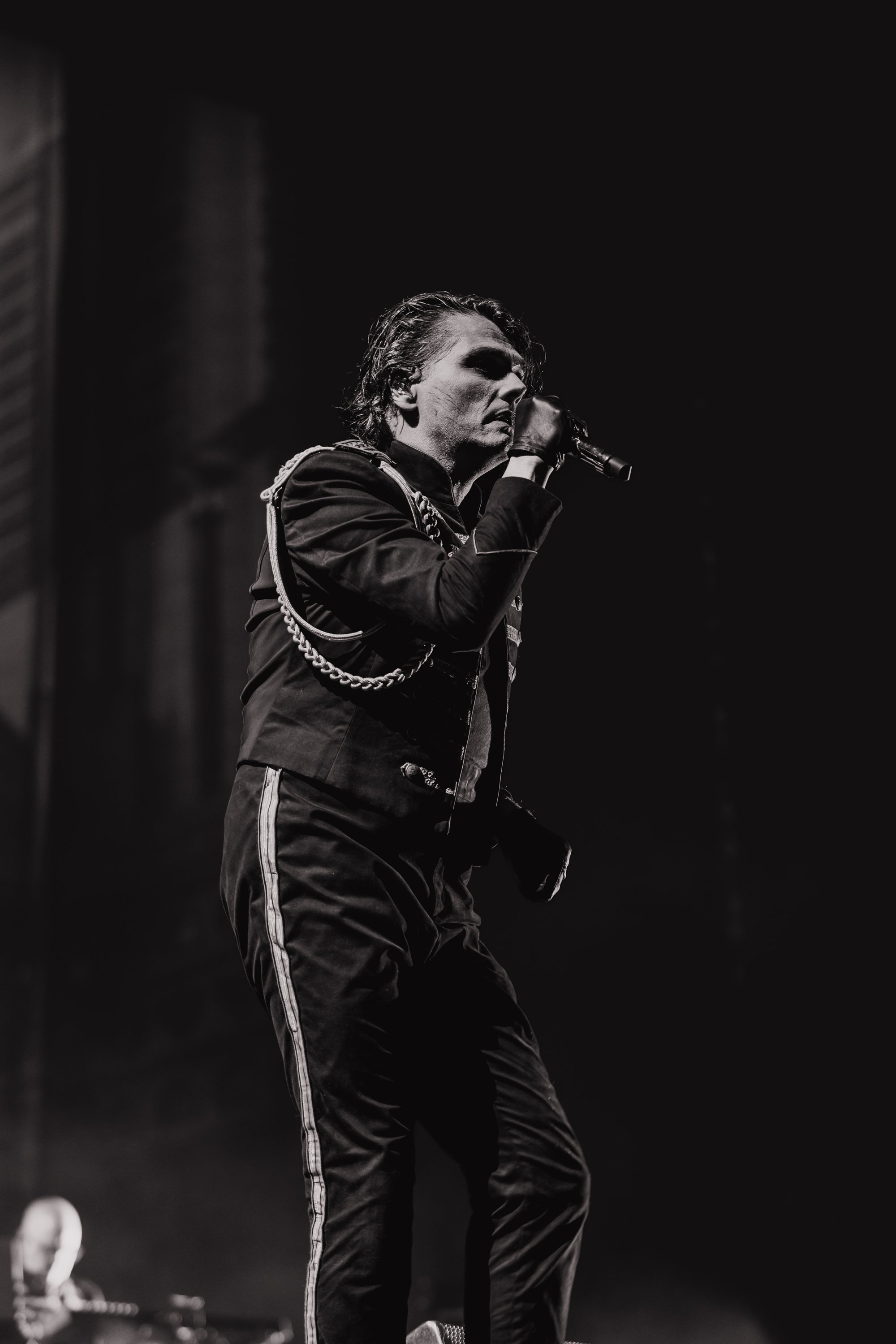 A black-and-white photo of a male singer performing on stage, holding a microphone close to his mouth and wearing a dark jacket with metallic embellishments, and dark pants with a stripe on the side.