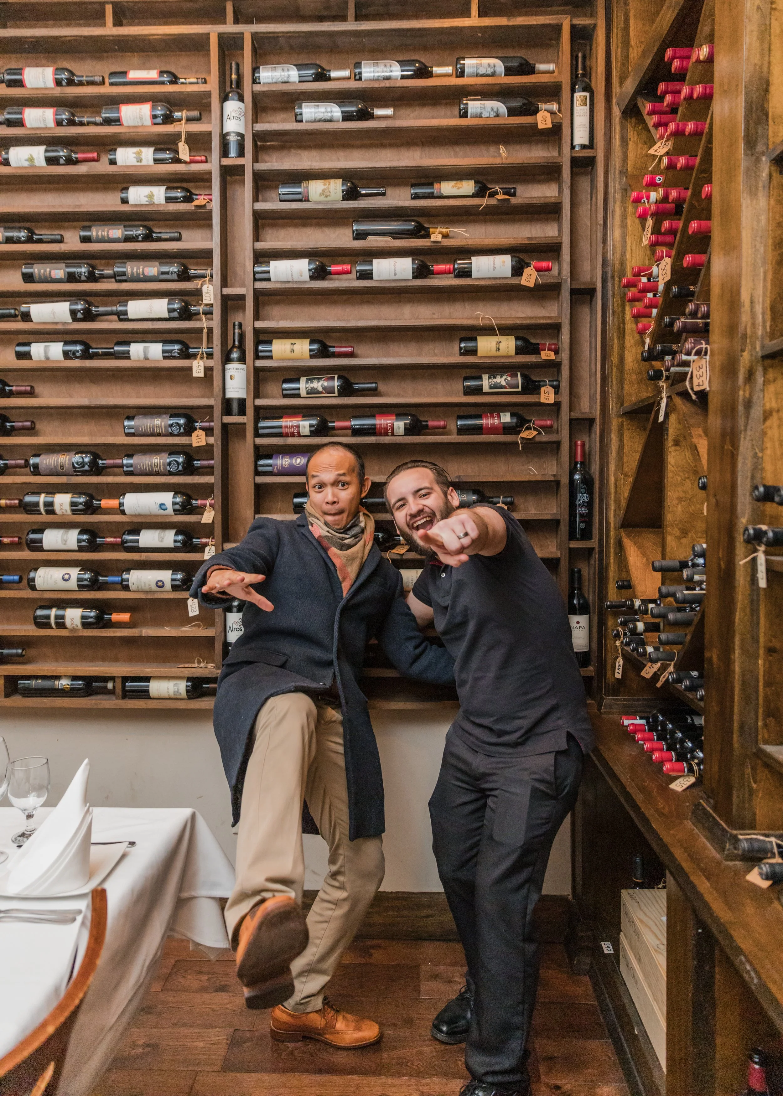 Two men posing playfully in front of a wine rack in a restaurant or winery setting, with smiles and one pointing at the camera.