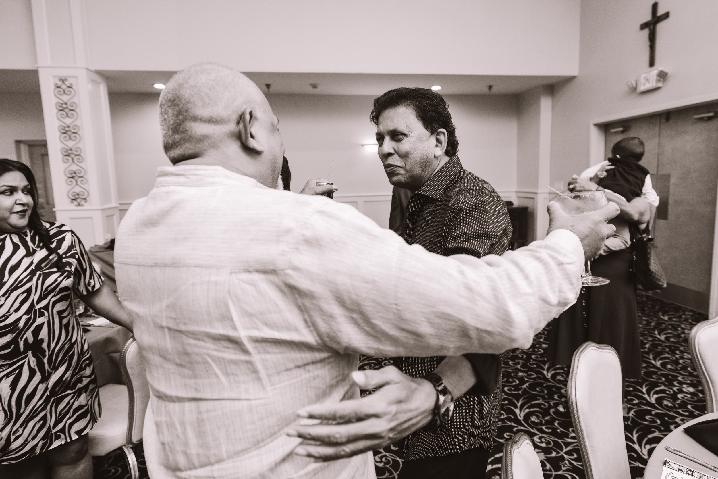 Group of people at social gathering in a banquet hall; two men engaged in conversation with drinks in hand, woman in patterned dress nearby, and a person hugging someone in the background.