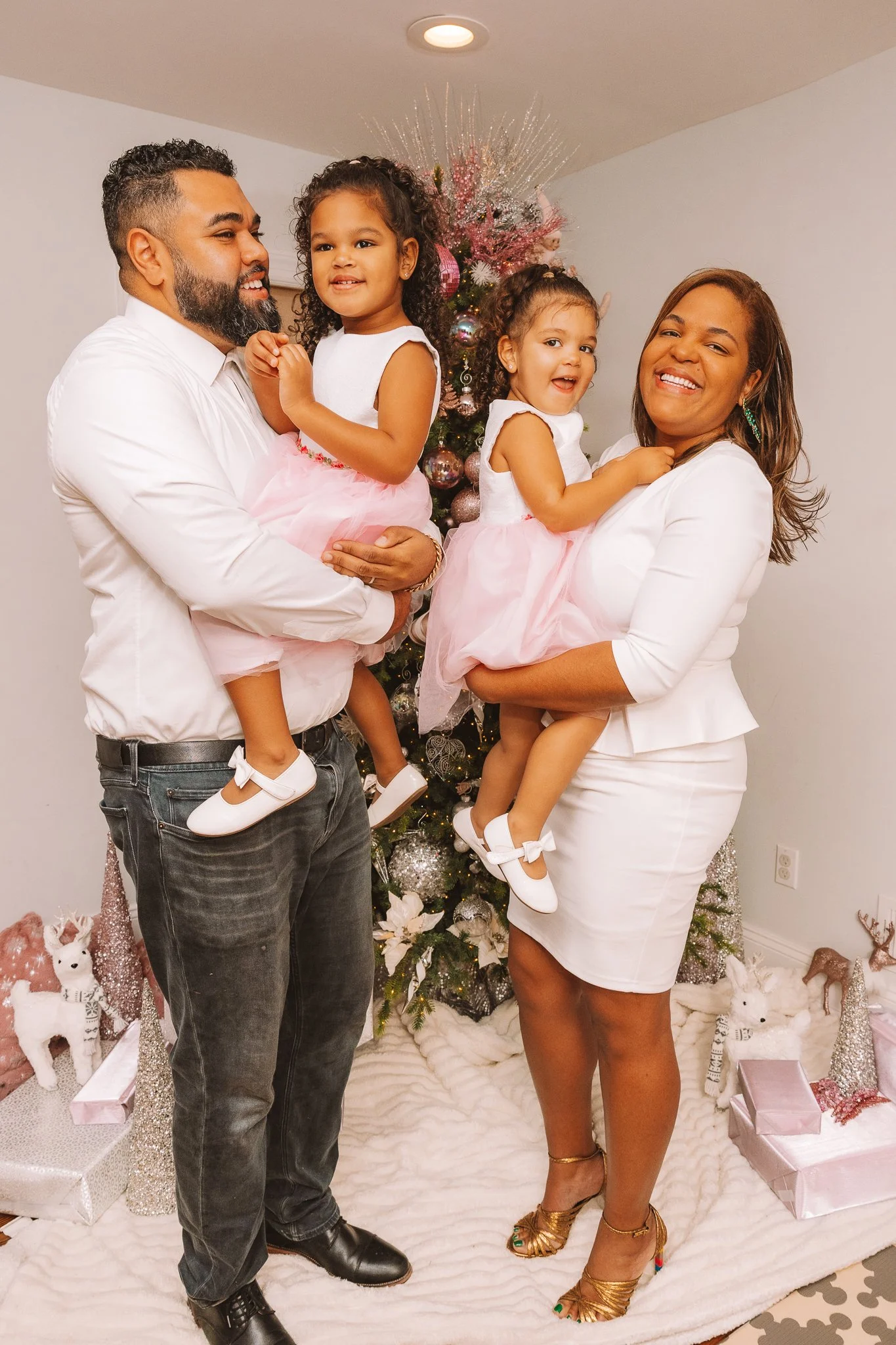 A family celebrating Christmas indoors, standing in front of a decorated Christmas tree. The man is holding a young girl, and the woman is holding a smiling girl. They are all dressed in white and pink outfits. Christmas decorations and presents are visible around them.