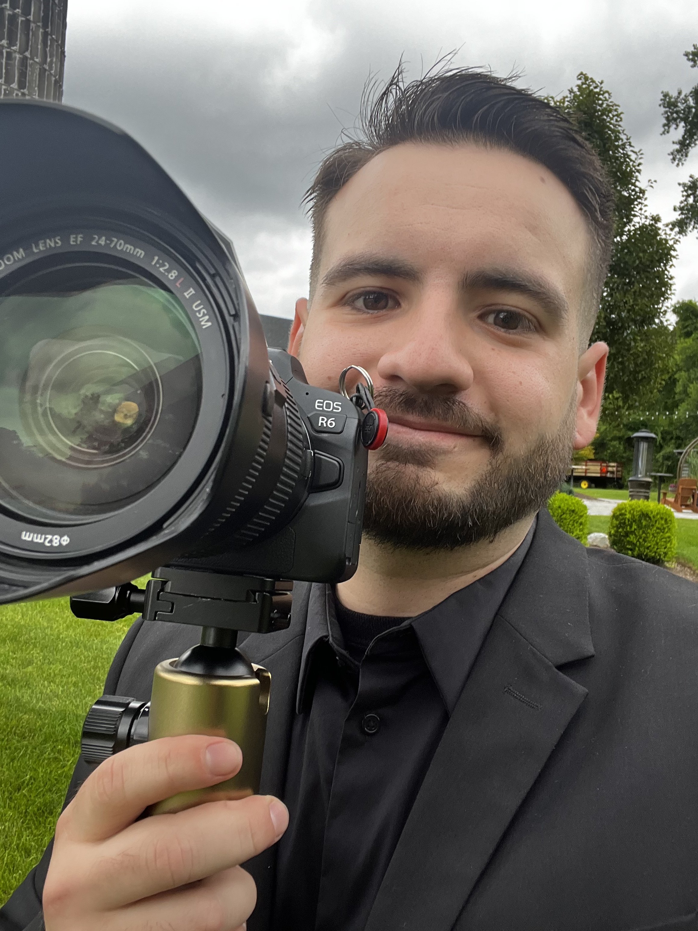 A young man with dark hair streaked with gray and a beard taking a selfie outdoors with a camera mounted on a tripod. The background features a green lawn, trees, and a cloudy sky.