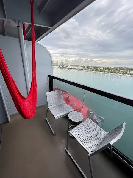 Cruise ship balcony with a red hammock chair, lounge seating, and a table overlooking a coastal city skyline and harbor.