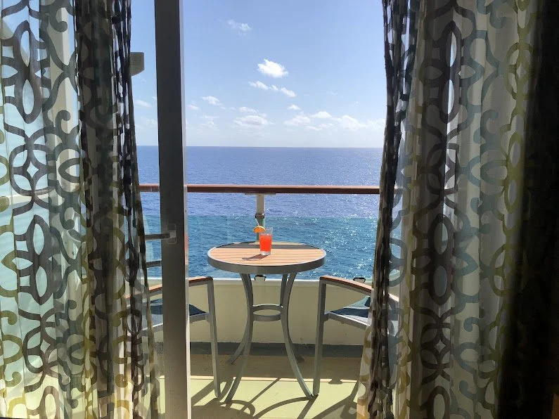 Ocean view through sheer curtains from a stateroom balcony, with a mai tai on the balcony table.