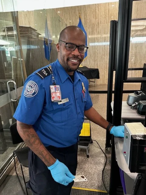 Security officer at TSA checkpoint smiling, wearing blue uniform and gloves, with a badge and glasses, standing next to a security scanner.