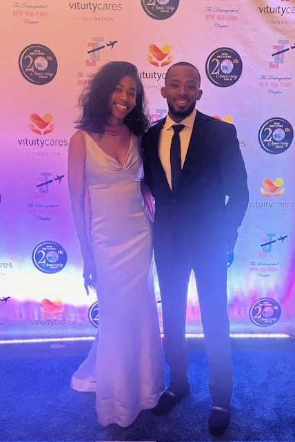A man in a black suit and yellow tie standing next to a woman in a white dress at an event with a step and repeat banner background.