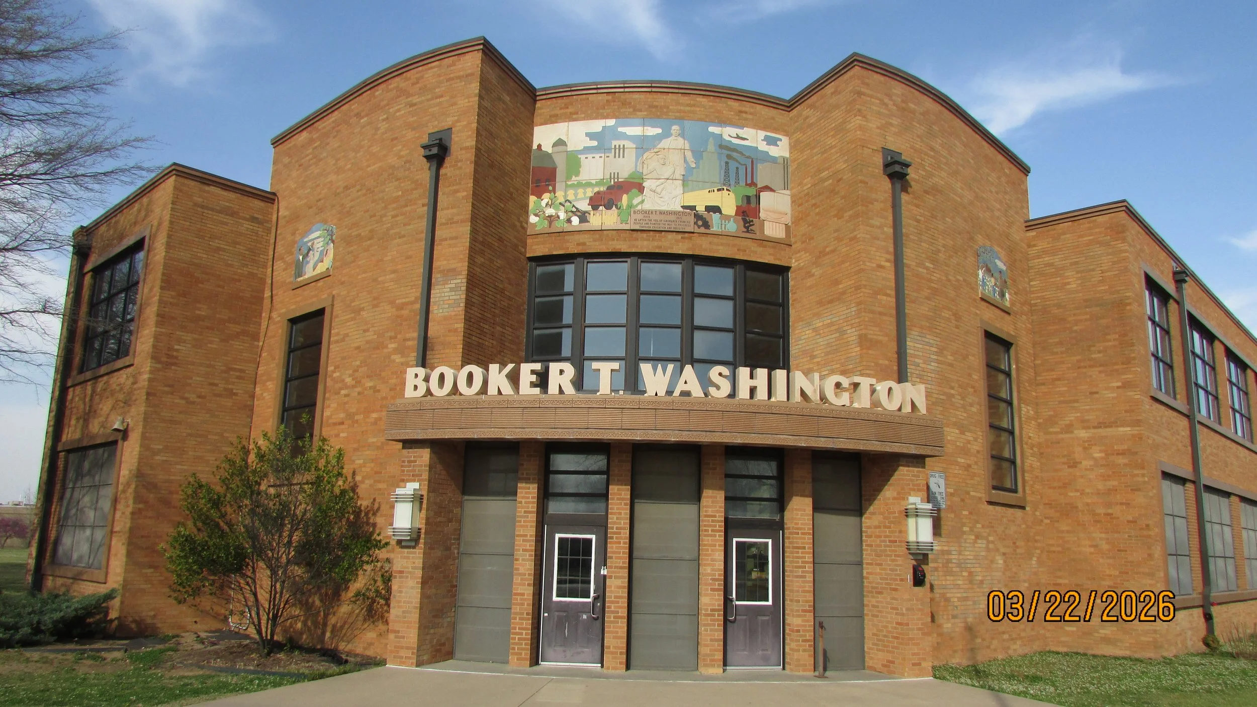 Front view of a brick building with bold white letters spelling Booker T. Washington, with decorative street lamps on either side of the entrance and a mural above the windows.