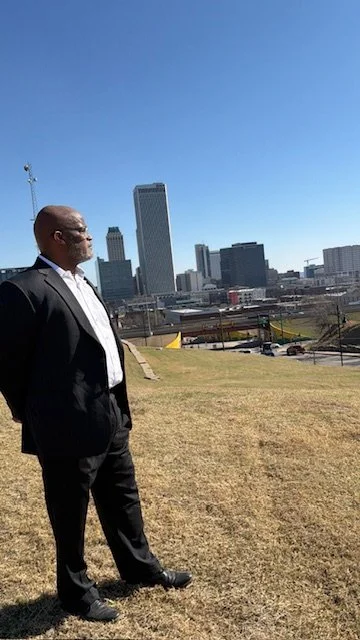A man in a suit standing on a grassy area with the city skyline in the background, including tall buildings and a clear blue sky.