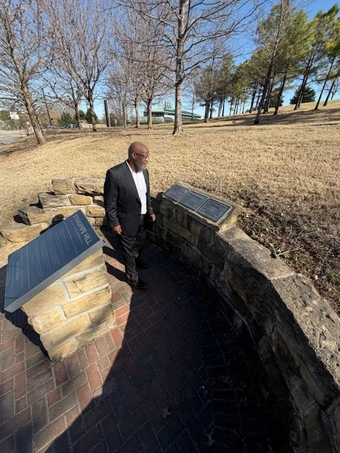 A man in a black suit and white shirt looking at historical plaques on a stone wall in a park with leafless trees and some green trees in the background.