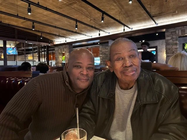 Two men sitting close together at a restaurant, smiling at the camera. One is wearing a brown sweater and the other a black leather jacket. There are people and TV screens in the background.
