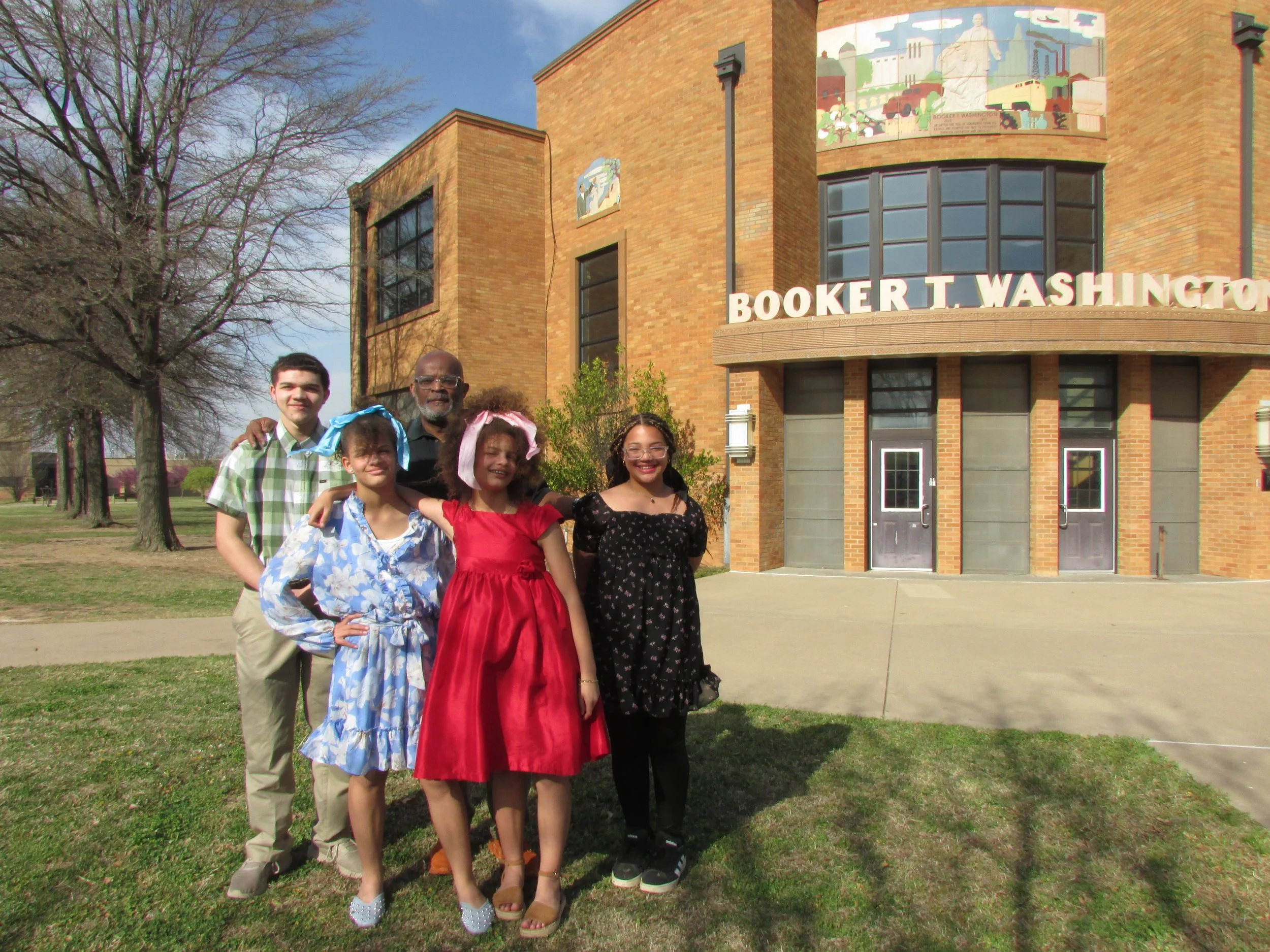 A group of five people, including children and an adult man, stands outside a brick building with the sign "Booker T. Washington" and a colorful mural above the entrance. The children are dressed in colorful, fancy clothes, some with headbands, and are smiling for the photo on a sunny day with a tree-lined area in the background.