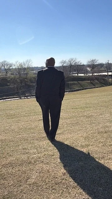 Man in a black suit walking on a grassy field during daytime, casting a long shadow, with trees and a river in the background.