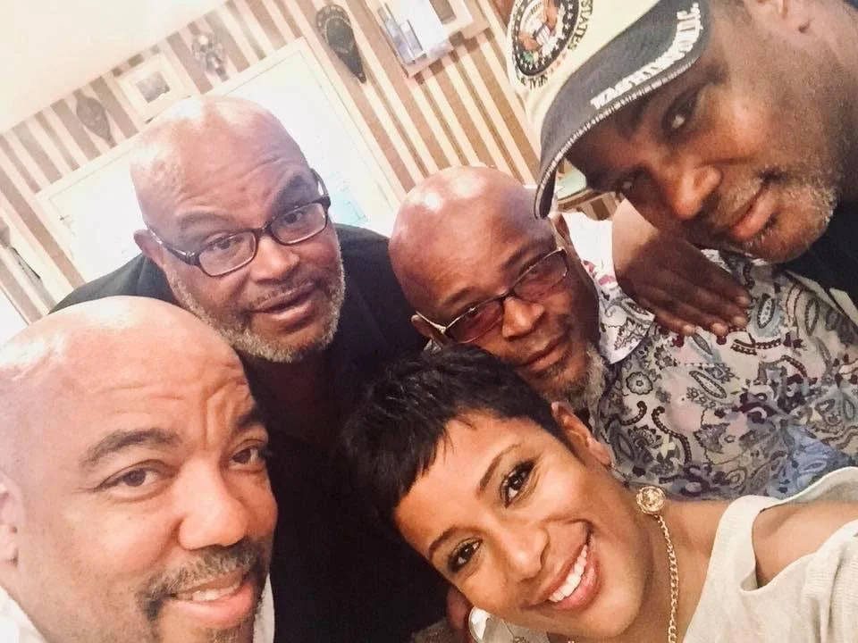 Five people taking a close-up group selfie indoors, four men and one woman, all smiling and looking at the camera.