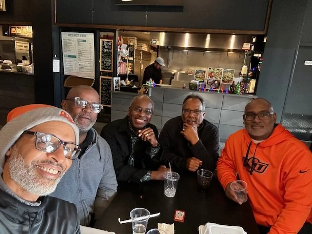 Five men sitting together at a table in a restaurant or cafe, smiling and posing for a photo. The man on the left wears glasses and a tan beanie, the second man wears glasses and a gray hoodie, the third man is smiling with glasses and a black jacket, the fourth man wears glasses and a black jacket, and the fifth man is in an orange hoodie.