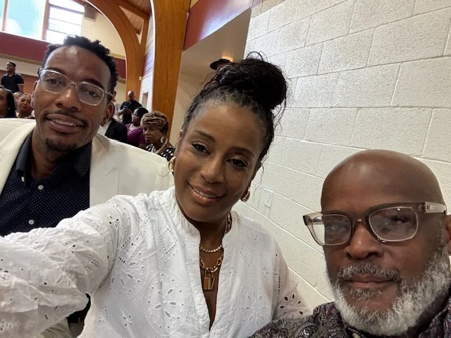 Three people taking a selfie indoors at an event, with people seated at tables in the background.