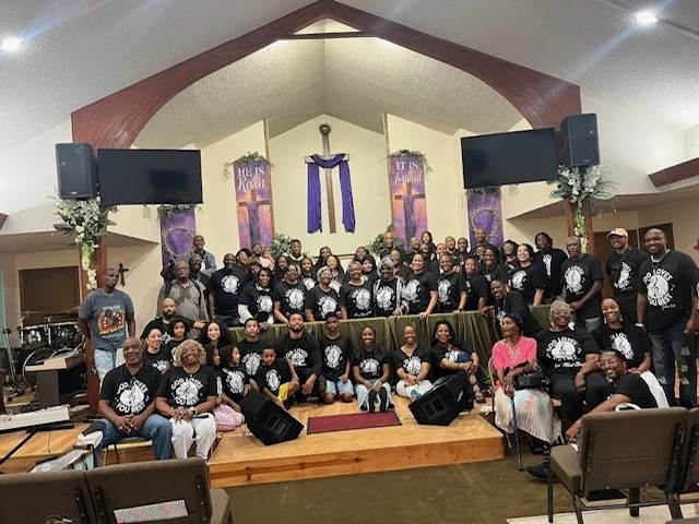 A large group of people gathered on the stage of a church or community hall, with a cross and religious banners in the background, and two large monitors on either side of the stage.